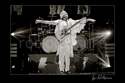 Horizontal black and white image of John 5 performing live on stage with an electric guitar, white stage outfit, outstretched arms, and bright backlights