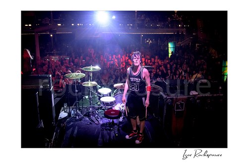 Horizontal color image of Zoltan Chaney standing beside his drum kit on stage with the audience visible behind him