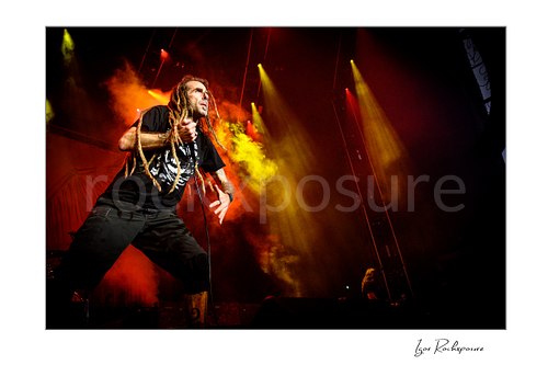 Horizontal color image of Randy Blythe of Lamb of God performing on stage with dreadlocks, handheld microphone, and red and yellow stage smoke