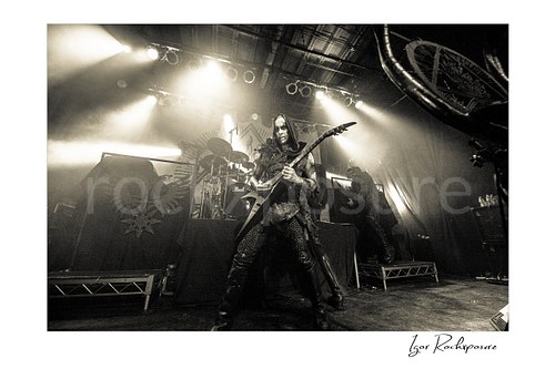 Horizontal black and white image of Nergal of Behemoth performing with guitar onstage with band members and bright overhead concert lights