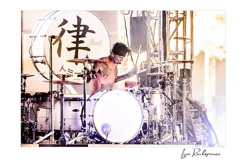 Horizontal color image of Tommy Lee performing at the drum kit during a Mötley Crüe concert, surrounded by cymbals and bright backlight