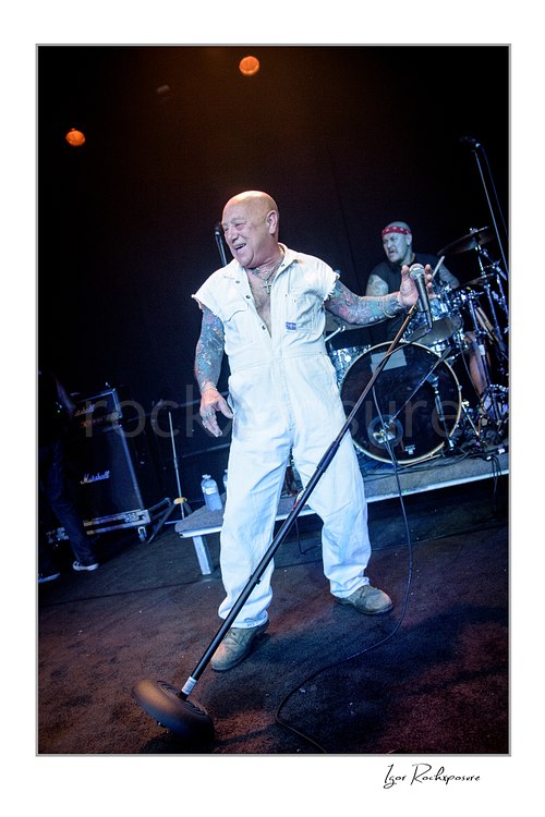 Vertical color image of Angry Anderson of Rose Tattoo performing with a microphone stand onstage with the drummer visible behind him