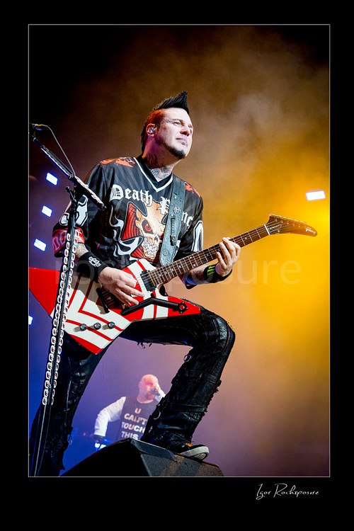 Vertical color image of Zoltan Bathory of Five Finger Death Punch playing guitar onstage under warm yellow concert haze