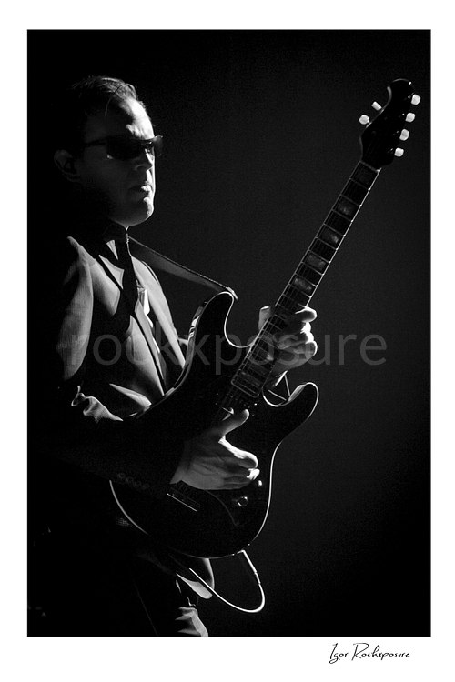 Vertical black and white image of Joe Bonamassa performing live on stage with his guitar in dramatic side lighting
