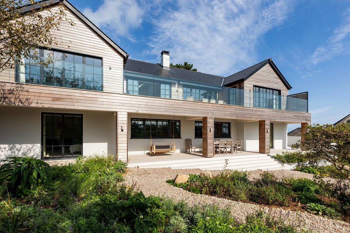 A modern two-story house in Cornwall with a wooden exterior, large windows, and a spacious balcony stands elegantly. Captured by an architecture photographer, the foreground reveals a lush garden with various plants adding to its charm.