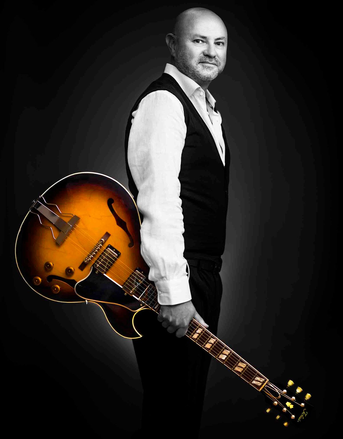 Portrait of Lonnie Pasco. He wears a white short, black vest. He is black and white and his guitar is in colour.