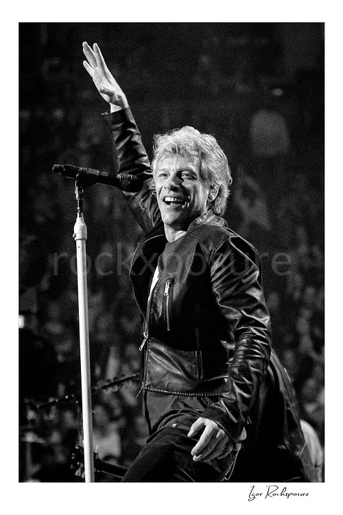Vertical black and white image of Jon Bon Jovi smiling on stage beside a microphone stand, with one arm raised during a live performance