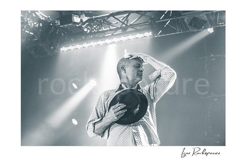Horizontal black and white image of Gord Downie of The Tragically Hip in an emotional live performance moment, holding his hat to his chest while pressing a white cloth to his forehead under soft stage light