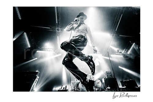 Horizontal black and white image of Gord Downie of The Tragically Hip captured high in the air off the stage in a dramatic low-angle wide-lens live performance shot, backlit by intense concert beams