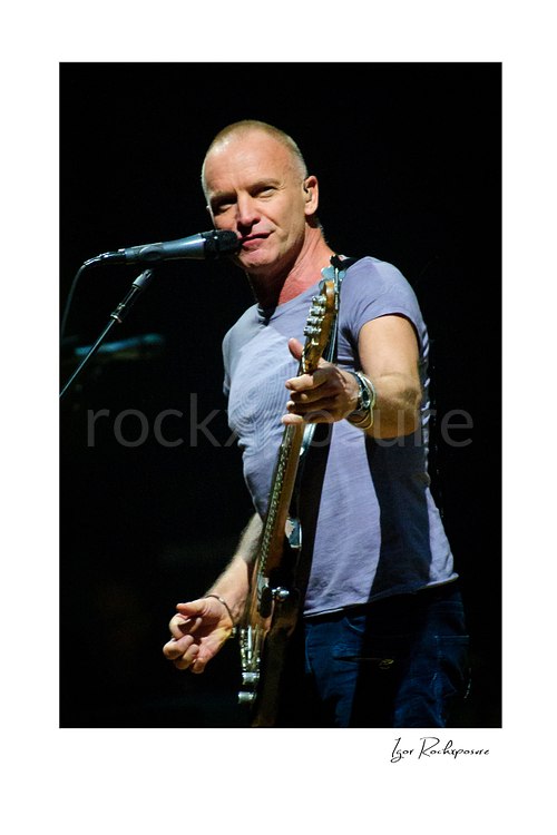 Vertical color image of Sting holding a bass guitar beside a microphone during a live performance