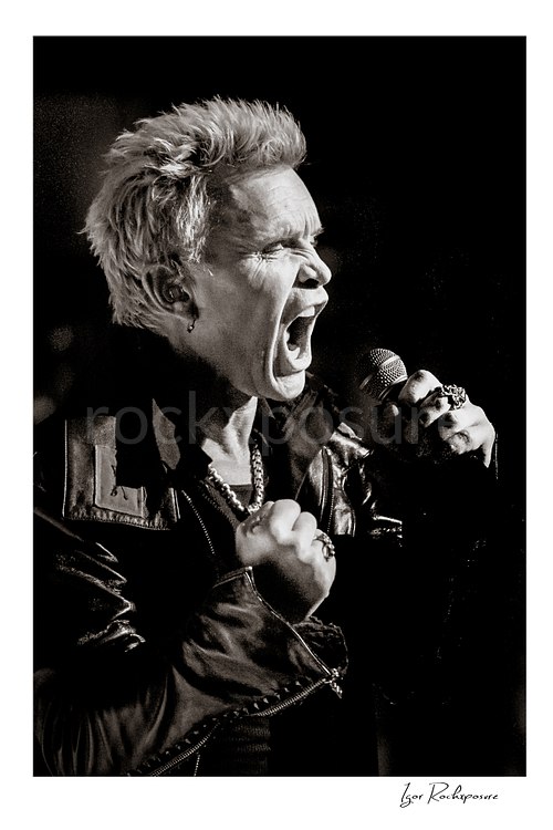 Vertical black and white image of Billy Idol singing into a microphone in a tight high-energy live performance close-up