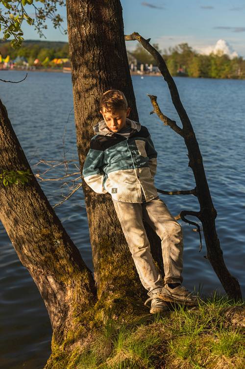Ein Junge in einer blau-beigen Cordjacke lehnt nachdenklich an einem Baumstamm direkt am Wasser im warmen Abendlicht.
