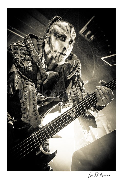 Vertical black and white image of Orion of Behemoth playing bass in an intense low-angle close-up with corpse paint and spiked stagewear