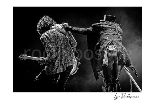 Horizontal black and white image of Steven Tyler and Joe Perry of Aerosmith performing live on stage from behind with glitter jackets, top hat, guitar, and drifting stage smoke
