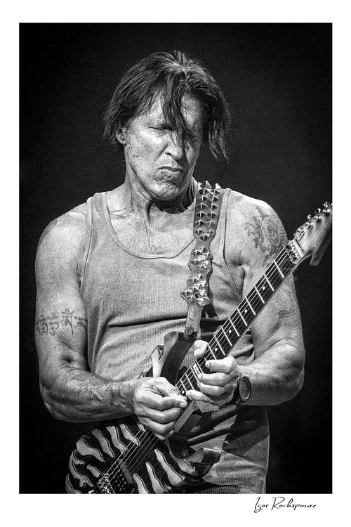 Vertical black and white image of George Lynch playing guitar on stage in a sleeveless shirt with a striped guitar