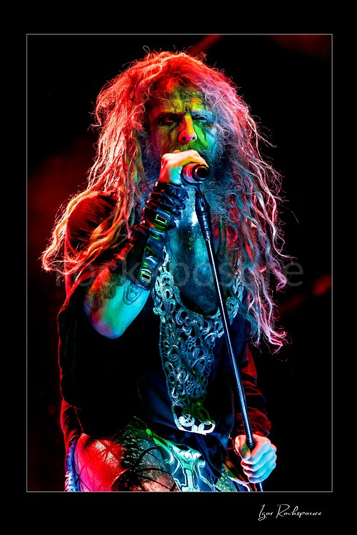Vertical color image of Rob Zombie performing live on stage with long dreadlocks, a handheld microphone, dramatic red and green lighting, and a black stage outfit