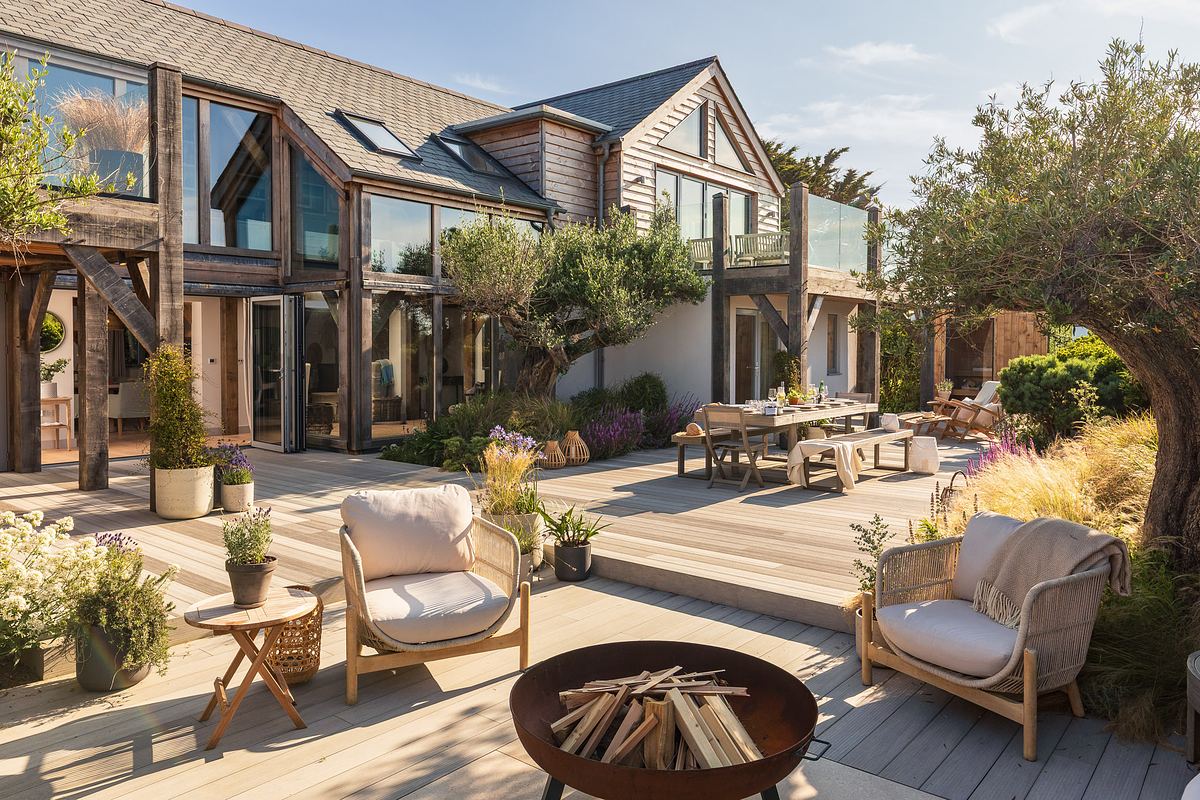 Modern two-story holiday let in Cornwall with large glass windows, wooden deck, outdoor seating, dining table, fire pit, and lush garden  architectural landscaping under a clear sky.