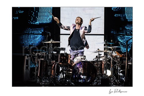 Horizontal color image of Dominic Howard of Muse standing at the drum kit with both drumsticks raised during a dramatic live performance