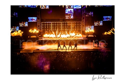 Horizontal color image of Trans Siberian Orchestra performing on stage with flames, smoke, and a packed arena crowd