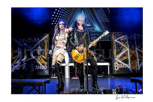 Horizontal color image of Calico Cooper and Chuck Garric of Beasto Blanco performing live on stage, with Cooper beside Garric at the microphone under blue stage light and the LED backdrop glowing behind them