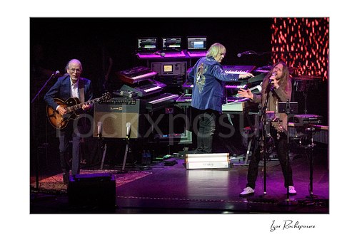 Horizontal color image of Steve Howe, Geoff Downes, and Jon Davison of Yes performing onstage together under purple concert lighting