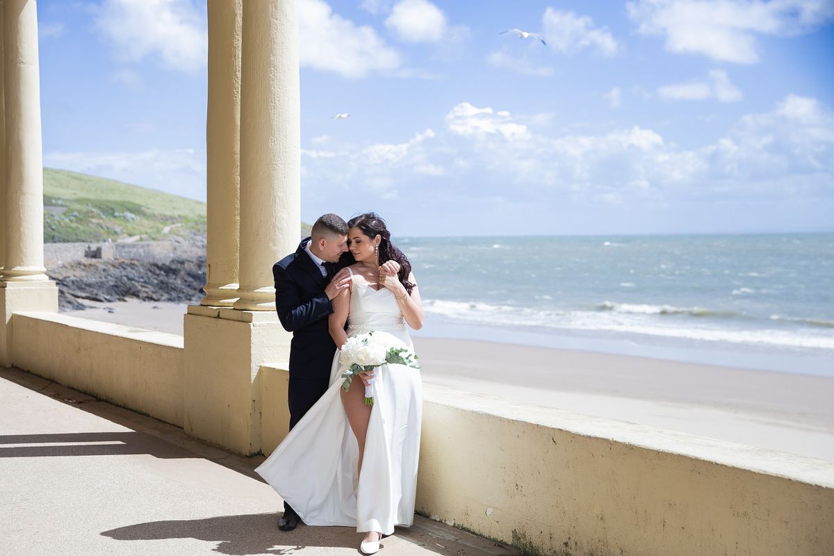 Bride and groom standing beneath seaside columns overlooking the beach at Barry Island, South Wales