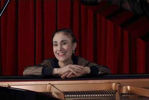 Myriam Birger at the piano in concert, leaning on the instrument in front of a red curtain