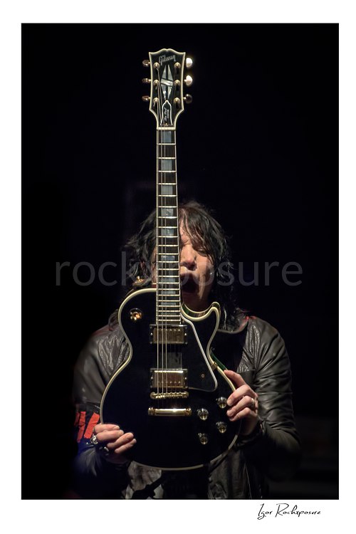 Vertical color image of Tom Keifer performing live on stage, holding a black guitar upright in front of his face against a dark background