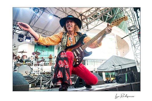 Horizontal color image of Randy Hansen kneeling on an outdoor ship deck stage with guitar and one arm outstretched during a live performance