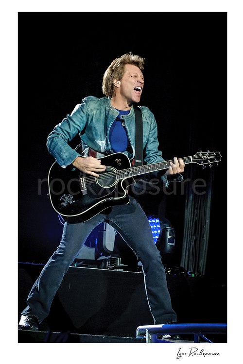 Vertical color image of Jon Bon Jovi performing live with a black acoustic guitar, singing on stage in a wide-legged stance with blue stage lights behind him