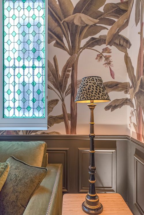 Détail d’une lampe avec abat-jour léopard devant une fenêtre vitrail et papier peint à motifs de palmiers dans un appartement haussmannien de 400 m² rue du Faubourg Saint-Honoré. Photographie d’architecture intérieure par Nicolas Karadimos.