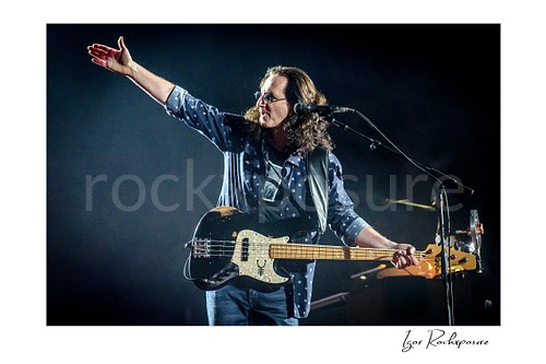 Horizontal color image of Geddy Lee performing with his bass guitar during a Rush concert, extending his arm beside a microphone under dramatic stage light