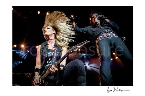 Horizontal color image of Nita Strauss and Alice Cooper performing live on stage with Nita Strauss playing guitar and her hair flying
