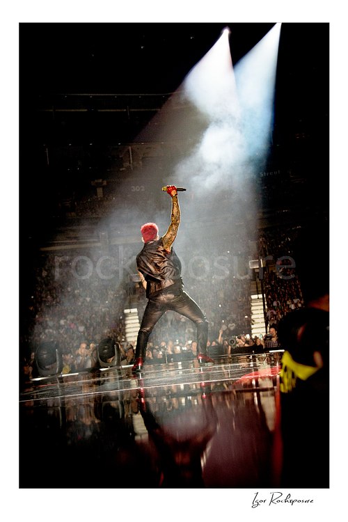 Vertical color image of Adam Lambert of Queen seen from behind onstage with one arm raised beneath a bright spotlight and drifting stage smoke