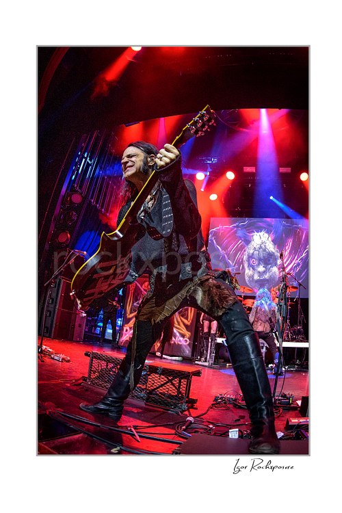 Vertical color image of Chuck Garric of Beasto Blanco playing bass on stage in a dramatic low-angle live shot, with vivid red lighting and the band backdrop visible behind him