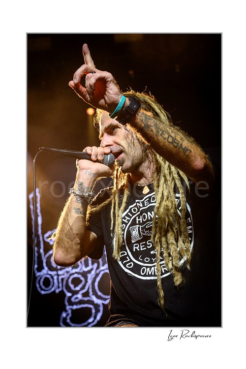Vertical color image of Randy Blythe of Lamb of God singing into a handheld microphone with raised finger and long dreadlocks