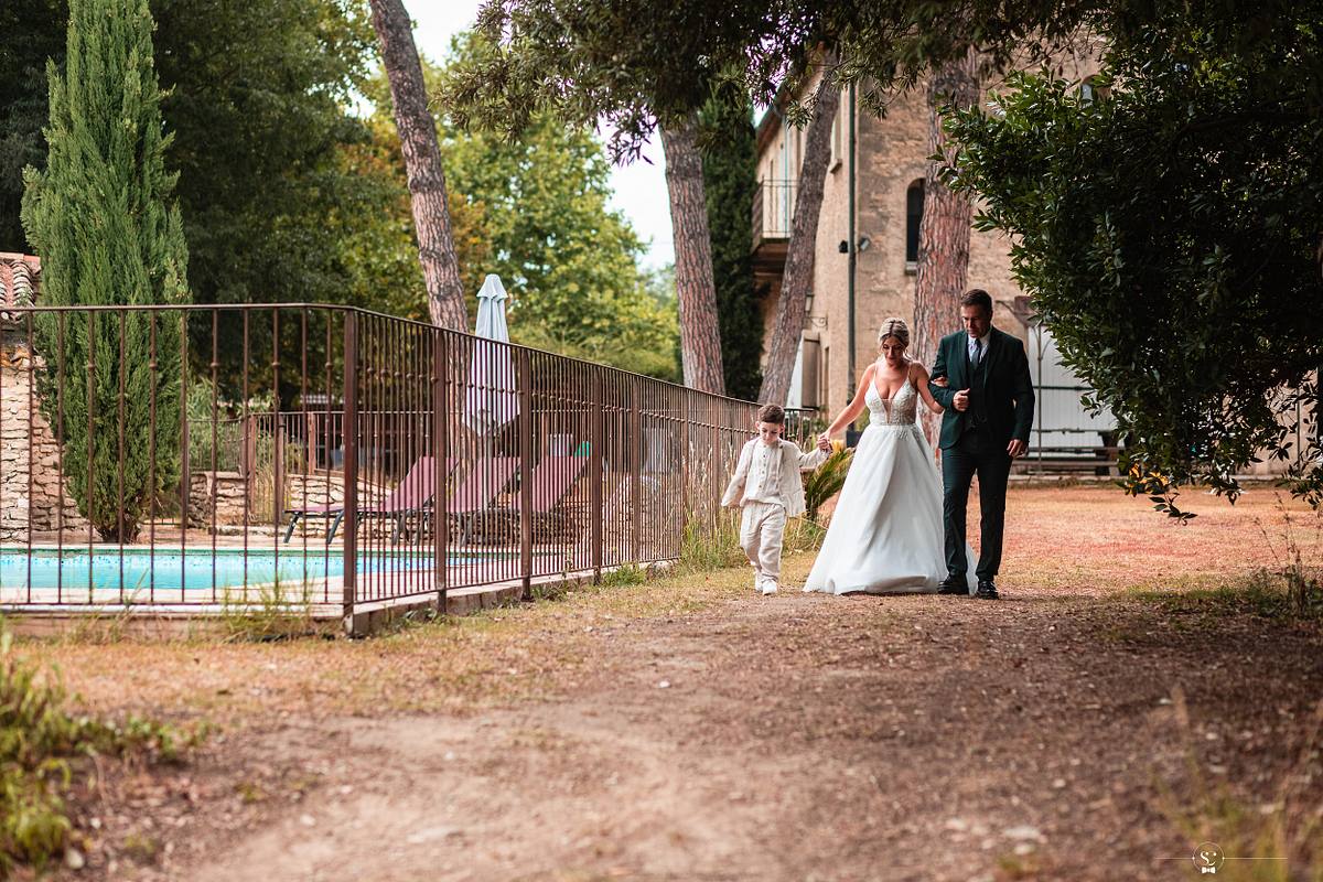 La mari&eacute;e en robe blanche et son p&egrave;re, &eacute;l&eacute;gamment v&ecirc;tu, marchent le long d'un sentier avec leur fils, avec en arri&egrave;re-plan une piscine et une architecture traditionnelle, capturant une promenade familiale intime avant la c&eacute;r&eacute;monie &agrave; N&icirc;mes