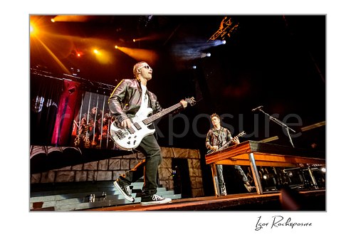 Horizontal color image of Johnny Christ and Synyster Gates of Avenged Sevenfold performing onstage under warm concert lighting beside a wooden stage riser