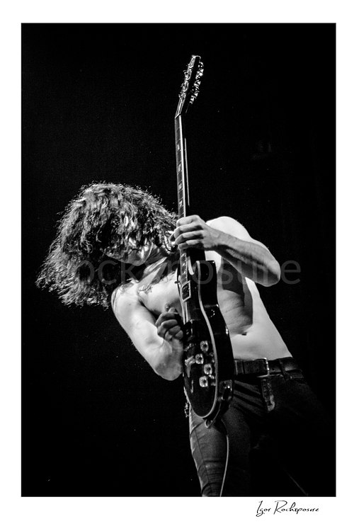 Vertical black and white image of Brent Woods playing guitar shirtless on stage with hair falling across his face