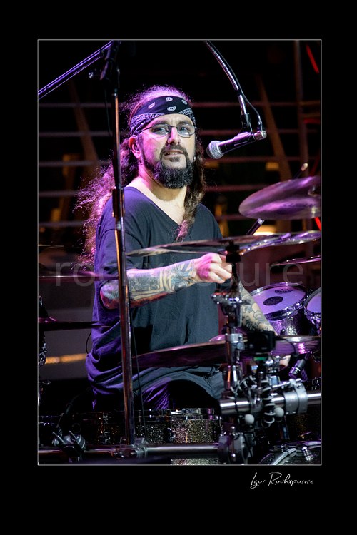 Vertical color image of Mike Portnoy at the drum kit looking toward the camera under purple stage lighting