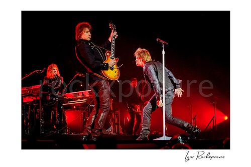 Horizontal color image of David Bryan, Richie Sambora, and Jon Bon Jovi of Bon Jovi performing live on stage under deep red lighting, with Jon Bon Jovi at the microphone