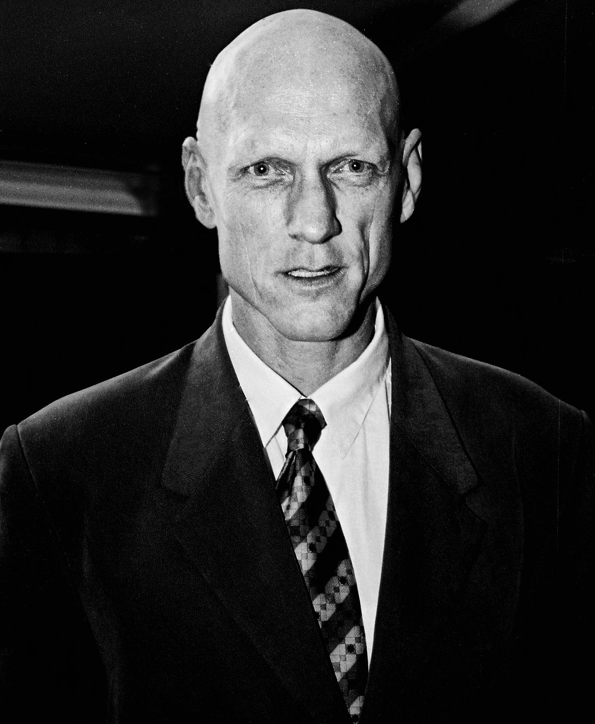 A portrait of singer, former Australian Federal Minister, Peter Garrett. He wears a black suit. Dark background.