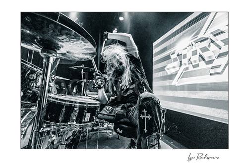 Horizontal black and white image of Robert Sweet of Stryper playing drums with raised drumsticks and flying hair