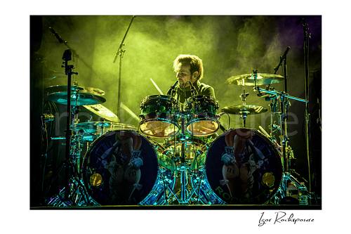 Horizontal color image of Rikki Rockett performing at the drum kit during a Poison concert, surrounded by cymbals and green stage smoke
