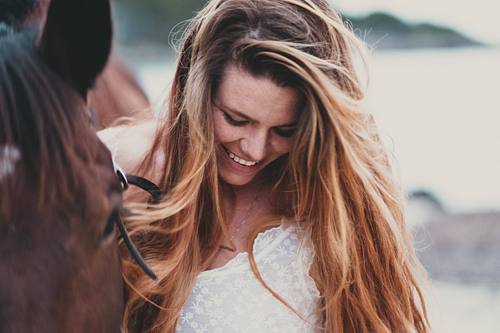Junge Frau lacht mit vom Wind verwehten Haaren neben braunem Pferd am See - emotionale Pferdefotografie by Carolin Kohlross in Amberg und Umgebung