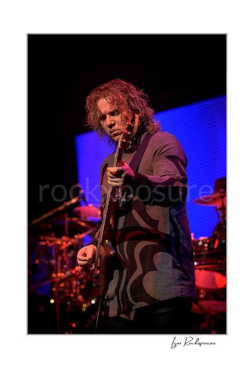 Vertical color image of Billy Sherwood of Yes playing bass onstage under red and blue concert lighting