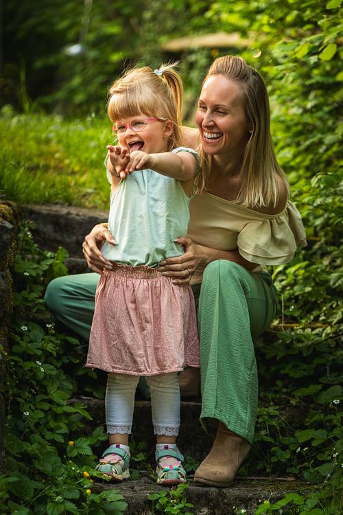 Eine Mutter hockt auf einer bewachsenen Steintreppe und hält ihre kleine, freudig lachende Tochter von hinten im Arm.