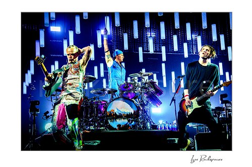 Chad Smith raises a drumstick above his purple kit between Flea holding a bass and Josh Klinghoffer playing guitar under glowing blue LED tubes.