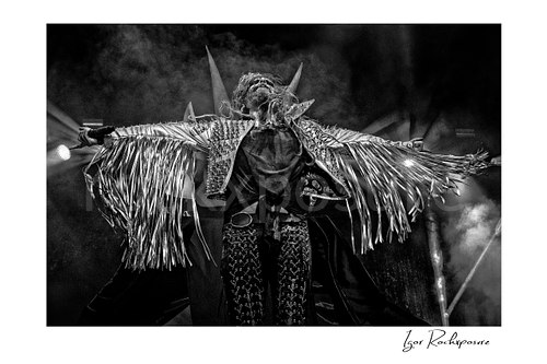 Horizontal black and white image of Rob Zombie performing live on stage with outstretched arms, a fringed jacket, stage smoke, and a horned backdrop