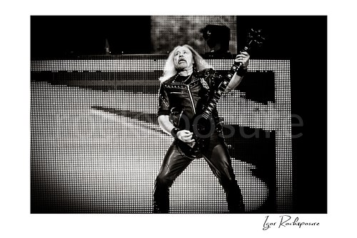 Horizontal black and white image of Glenn Tipton of Judas Priest performing live on stage, playing electric guitar in a wide stance against a bright LED screen backdrop